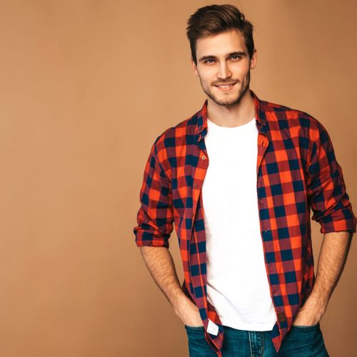Portrait of handsome smiling stylish hipster lumbersexual businessman model dressed in red checkered shirt. Fashion man posing on golden background