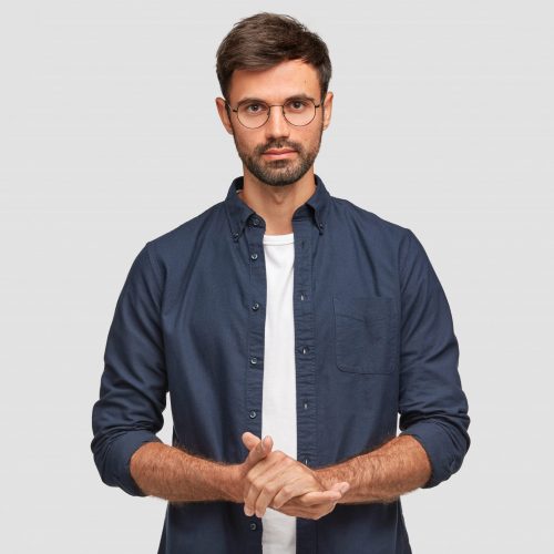 Waist up portrait of handsome serious unshaven male keeps hands together, dressed in dark blue shirt, has talk with interlocutor, stands against white background. Self confident man freelancer